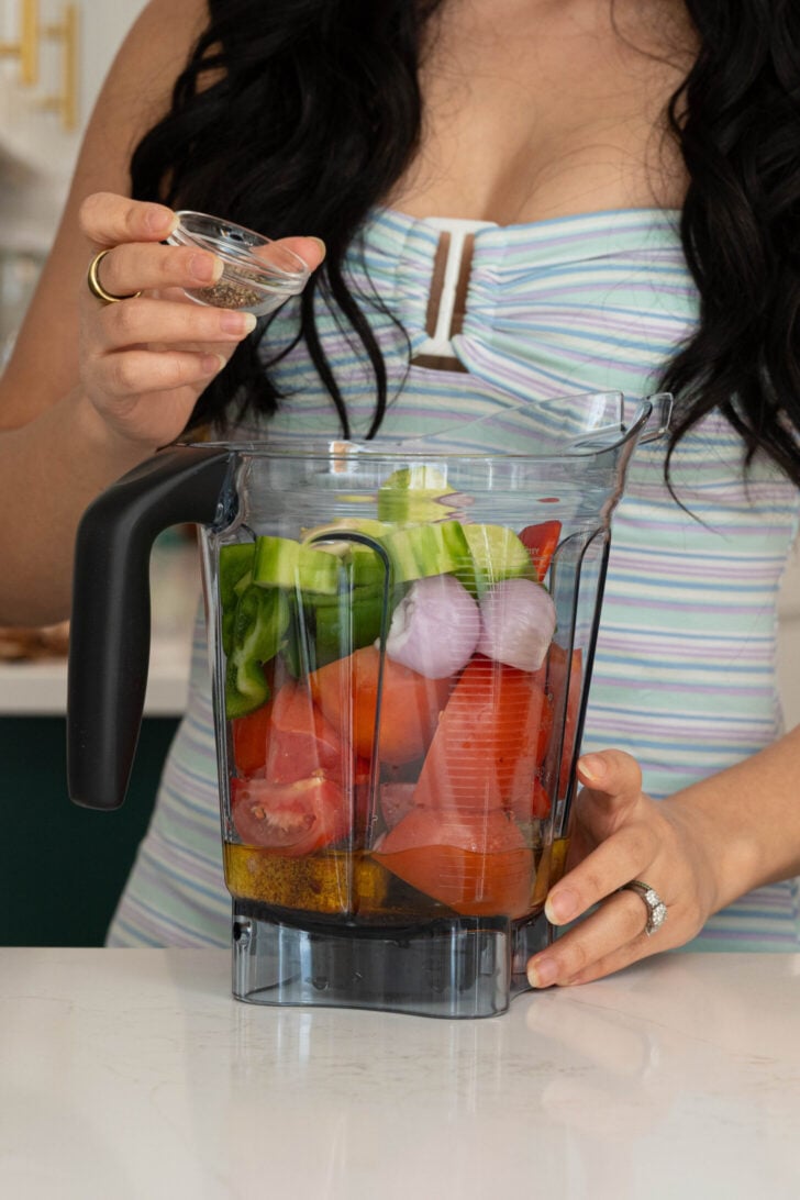 A girl adding pepper to a blender.